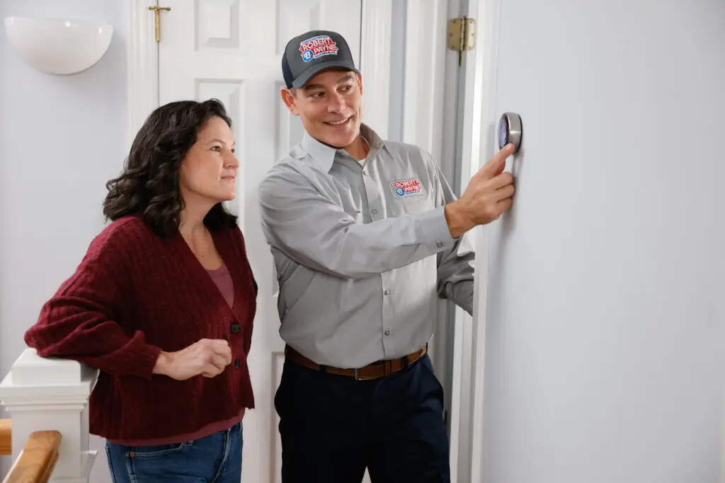Technician showing homeowner how to use her new smart thermostat on a white wall in a hallway.