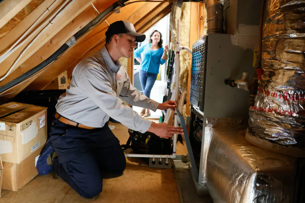 Technician servicing an HVAC system in a home's attic. Ductwork visible. Homeowner watching from open doorway in background.